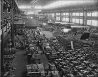 Interior view of a factory floor with dozens of tractors and machines stretching to a point in the background, with windows emitting light at the back and at the sides.