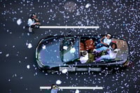 Two women, viewed from overhead, ride in a black convertible during a parade.