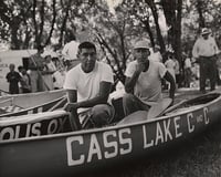 Two Ojibwe men crouch in a canoe with the words "Cass Lake" written on it in white capital letters.