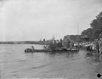 People crowd the shore of a lake and a dock stretching into open water.