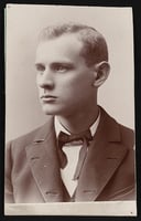 Photograph of 3/4 view of a man. He is wearing a blazer with a wide lapel, a ribbon bowtie, and a shirt with a white collar,