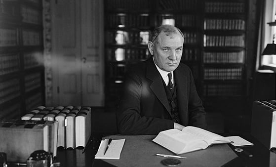 A man wearing a suit jacket and tie sits in a study with a book open on the desk in front of him.