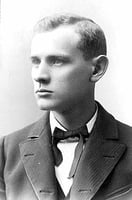 Photograph of 3/4 view of a man. He is wearing a blazer with a wide lapel, a ribbon bowtie, and a shirt with a white collar