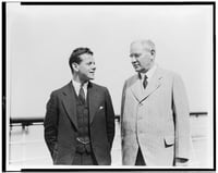 A young man in a dark suit and tie stands next to an older man in a light-colored blazer and glasses. The two men are looking at each other.