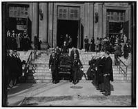 Eight men in formal attire carry a flower-laden casket down a set of stairs. They are surrounded by rows of people in dark formal attire.