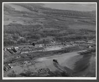 A bird's-eye view of a sprawling factory with buildings arranged into a narrow rectangle, with wooded land in the background.
