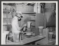 A worker wearing a brimmed cap and a respirator mask sprays glaze on a ceramic vase.