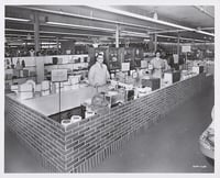 Two workers stand at sales counters behind a faux-brick wall.