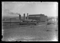 A five-story brick building sits at center with utility buildings to the right and smokestacks to the left.