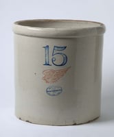 A dove-gray ceramic cylinder with the numeral 15 and a red wing image stamped on it sits in front of a lighter gray background.