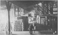 A worker in the foreground pushes a wheelbarrow down an aisle that leads between pyramidal stacks of ceramic containers.