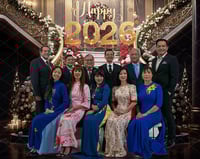 Two rows of people, one seated and one standing, are superimposed over a background featuring a staircase and the words "Happy 2026."