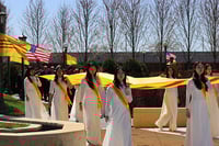 Nine women (visible) in long white dresses with yellow and red sashes carry a yellow and red flag.