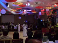 A woman in white traditional Vietnamese dress stands alone on a dance floor as a band performs behind her (right) and an audience looks on (background and foreground).