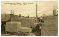 Stacks of wooden boards of varying heights sit together in a lumberyard, with an elevated track to the right.