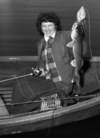 A woman sitting in a fishing boat holds a fishing line in her right hand and a caught fish in her left.