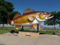 Yellow-and-brown sculpture of a fish with a white belly supported on a platform in front of trees and a blue sky.