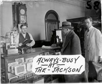 Image from a glass plate negative showing the front desk of the Jackson Hotel, ca. 1926. Cigars for sale are visible in the case, and a timetable for the Anoka Electric Line is fixed to the back of the cash register. Art Anderson is one of the men in the photograph. This image was used as an advertisement at Green’s Theater, located on Main Street in Anoka a few blocks from the hotel. Photographer unknown. Used with the permission of the Anoka County