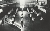 Black and white photograph of girls in uniform performing calisthenics during a physical education class on the second floor of Kiehle Hall, 1927.