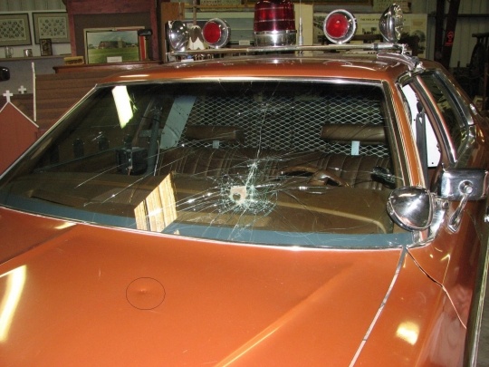 The cracked windshield and dented hood of Val Johnson's 1977 Ford LTD squad car, preserved unrepaired at the Marshall County Historical Society. Ford windshield specialist Meridan French could find no conventional explanation for the inward-and-outward fracture pattern.