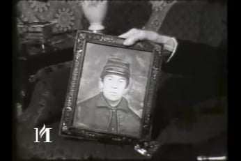 A man holds a painting of himself with his left hand while sitting down in a chair.