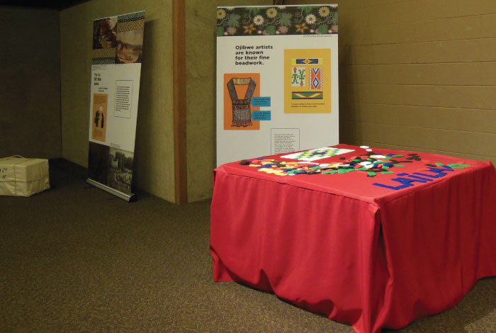 A table with red tablecloth holds tiles of multiple colors in front of a sign for beadwork designs.