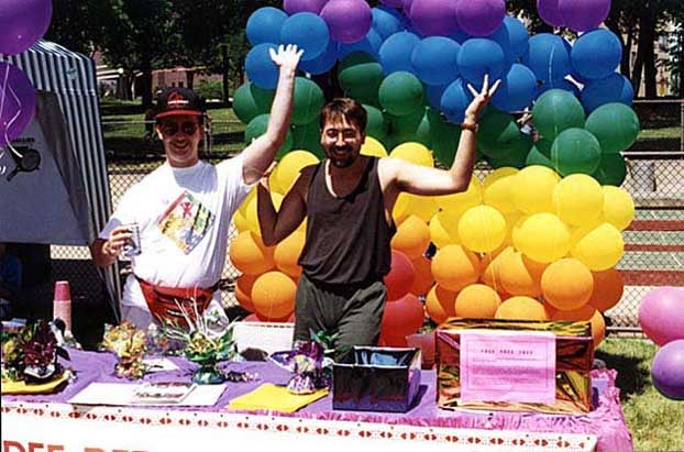 Two men stand in front of a rainbow of balloons behind a booth.