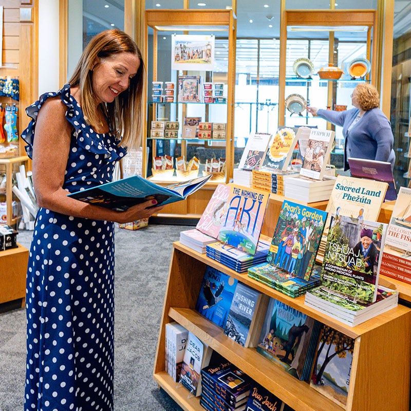 A smiling woman in a blue and white polka dot dress reads an open book in a brightly lit gift shop. Next to her is a wooden display table showcasing various books, while another shopper browses shelves in the background.