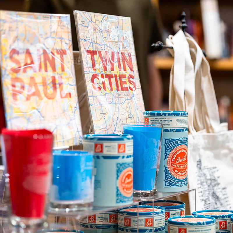 A retail display featuring packaged street maps of Saint Paul and the Twin Cities, alongside red and blue custom engraved pint glasses and a white canvas tote bag.
