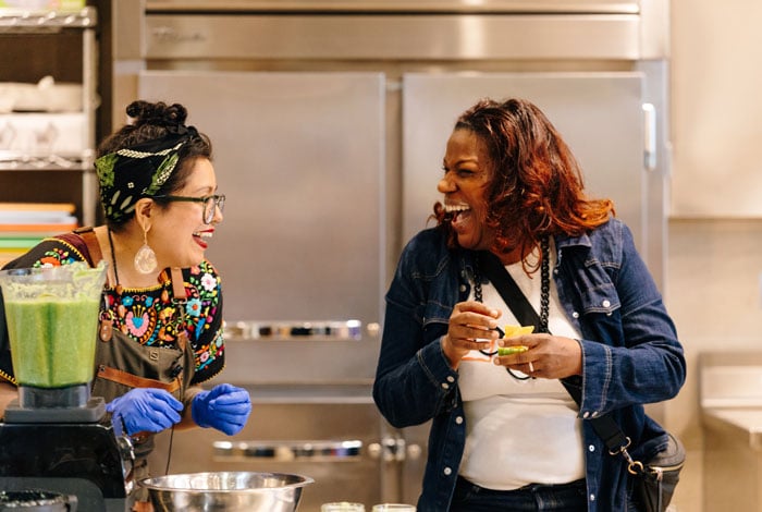 A Taste of History immersive culinary workshop exploring identity and foodways. Two women laugh joyously in a kitchen. A chef in an embroidered top, apron, and blue gloves stands by a blender of green sauce, sharing a fun storytelling moment with a laughing participant in a denim jacket. A Taste of History immersive culinary workshop exploring identity and foodways. Two women laugh joyously in a kitchen. A chef in an embroidered top, apron, and blue gloves stands by a blender of green sauce, sharing a fun storytelling moment with a laughing participant in a denim jacket.