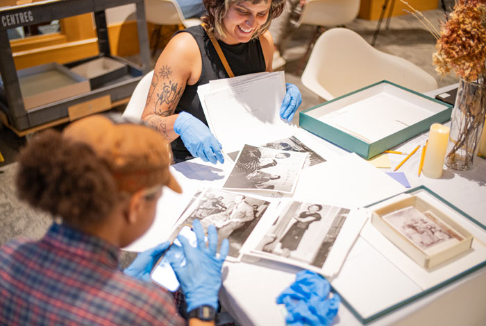 An Artist & Archives interactive workshop activating community history. Two people wearing blue protective gloves smile while examining historic black-and-white photographs spread across a table. One person uses a smartphone to take a picture of the archival materials. An Artist & Archives interactive workshop activating community history. Two people wearing blue protective gloves smile while examining historic black-and-white photographs spread across a table. One person uses a smartphone to take a picture of the archival materials.