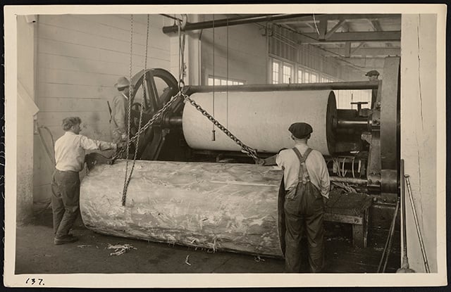Workers inside a lumber mill operate a roller machine.