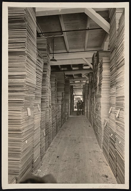 Stacks of milled lumber inside a lumber mill, with stacks piled up on either side of an empty walkway