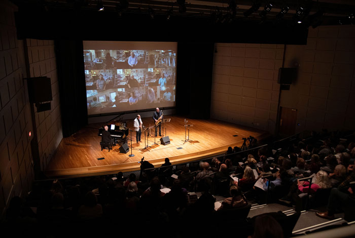 A History on Screen event sharing inclusive histories. An audience in a dark auditorium watches three musicians perform on a lit stage with a piano and woodwinds. Behind them, a large projection screen shows a grid of video participants, blending live performance with community dialogue. A History on Screen event sharing inclusive histories. An audience in a dark auditorium watches three musicians perform on a lit stage with a piano and woodwinds. Behind them, a large projection screen shows a grid of video participants, blending live performance with community dialogue.