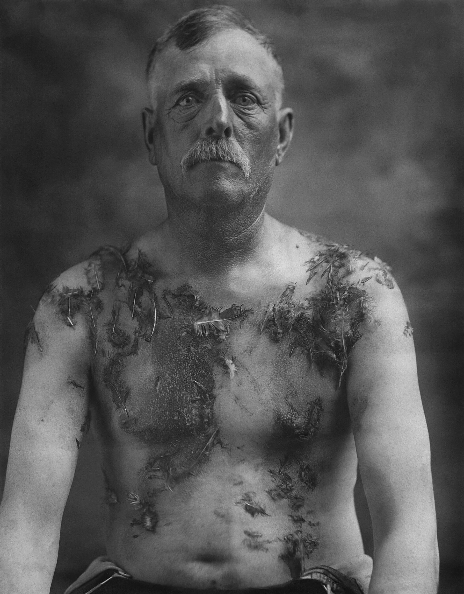 Photograph of a man facing the camera directly. His chest and shoulders are coated with tar and feathers.