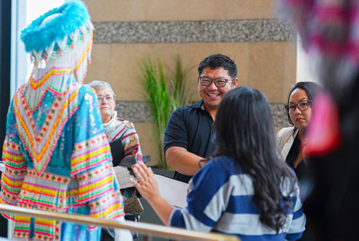 A Legacy Partnership event for the Hmong 50th. A smiling man engages in lively conversation with a small group. In the foreground stands a figure in vibrant traditional Hmong attire, featuring a bright blue headdress and intricate colorful embroidery, sharing cultural history. A Legacy Partnership event for the Hmong 50th. A smiling man engages in lively conversation with a small group. In the foreground stands a figure in vibrant traditional Hmong attire, featuring a bright blue headdress and intricate colorful embroidery, sharing cultural history.