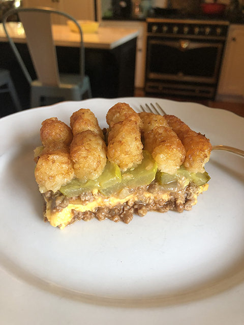 A serving of tater tot hotdish (showing its layers of ground meat, cheese, pickles, and tater tots) sits atop a white plate, with a kitchen scene in the background.