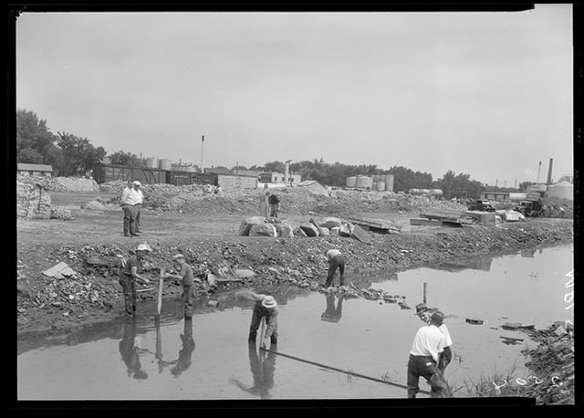 Black and white photograph of a construction site along a creek. Six workers lay wood, stone, and stakes in the creek, while two onlookers watch from the land next to the water.