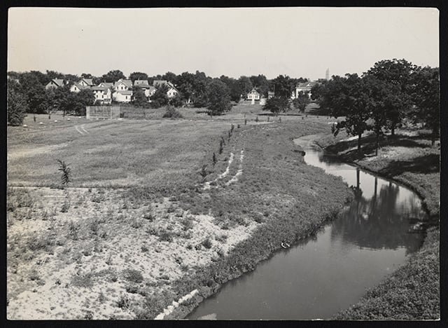 Lightly sepia-toned black and white photograph of Ȟaȟá Wakpádaŋ (Bassett Creek) curving through a field with a baseball diamond. White houses and tall, dark trees line the background.