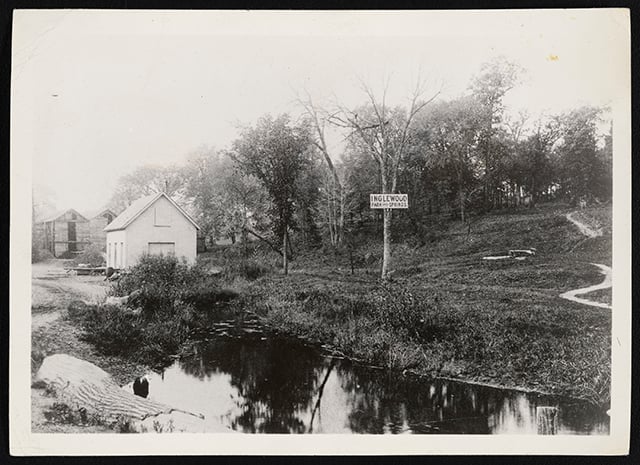 Black-and-white photograph of a spring at Bassett Creek in the foreground and a sign that reads “Inglewood Park and Springs.” Behind the creek is a white wooden house and a grassy hill with trees and a trail.