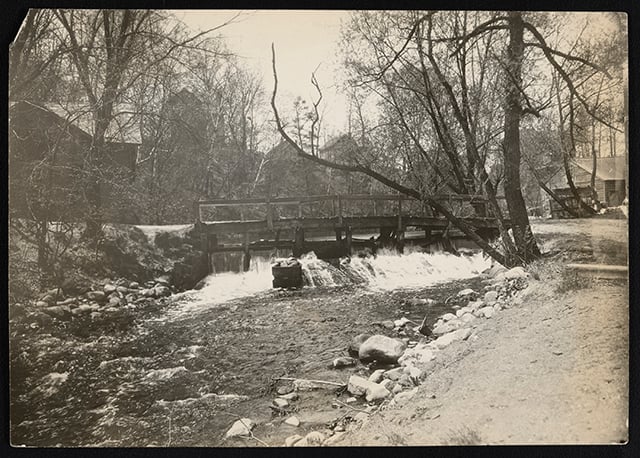 Lightly sepia-toned photograph showing a wooden dam in Bassett Creek. Around the creek and dam are dirt paths, bare trees and wooden houses in the background.