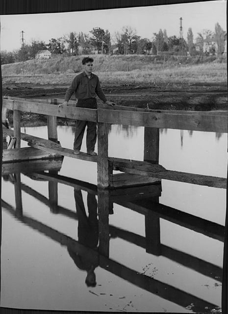 A black-and-white photograph depicting a young man walking across a bridge over Bassett Creek. The bridge is made of single, connected wood planks along with a rough wood handrail. 