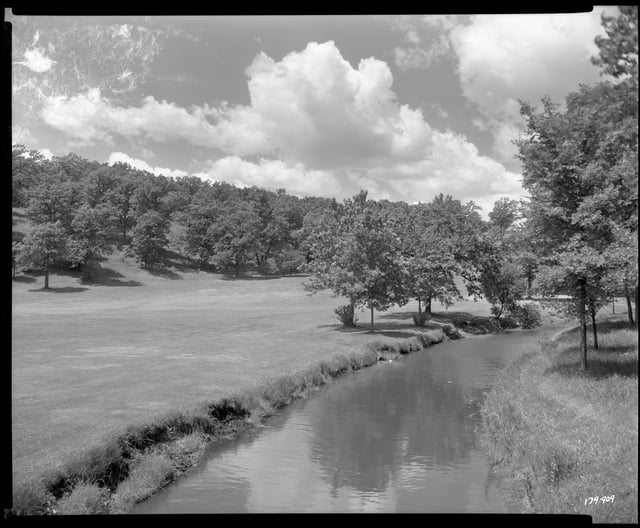Black-and-white photograph of a glassy portion of Bassett Creek curving inside Theodore Wirth Park against a grass field. Trees line the creek and the background against a cloudy sky. 