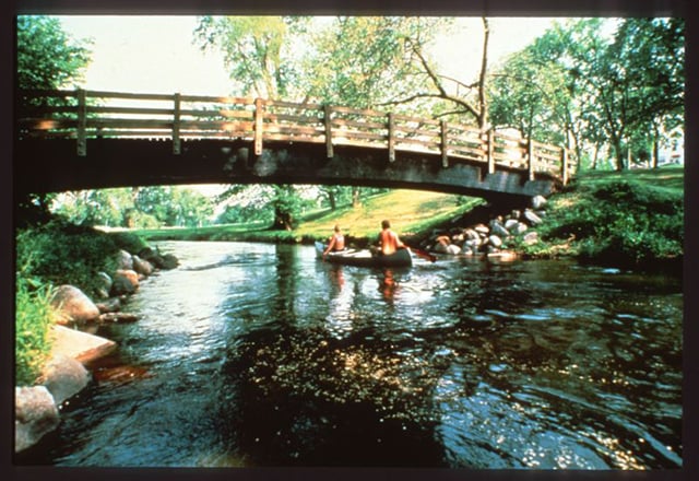 Color photograph of a couple paddling a canoe through a creek, heading under a wooden bridge. Grassy shores line the river with rocks underneath the bridge’s entrances.