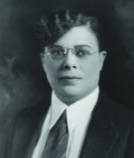 The head and shoulders of a woman with permed, side-swept bangs, wearing a suit and tie.