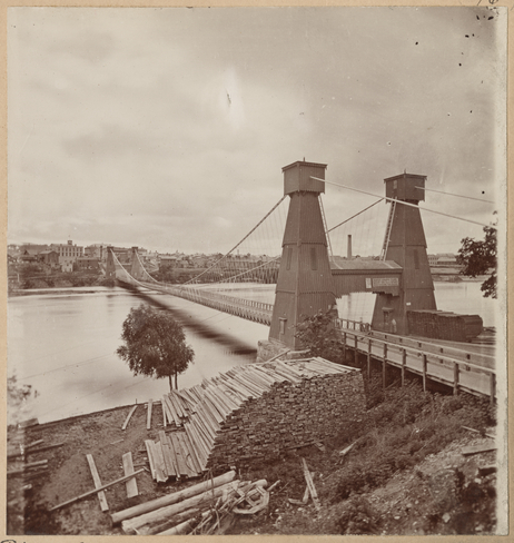 Sepia photograph of a suspension bridge. Stacks of logs are piled up in the foreground.
