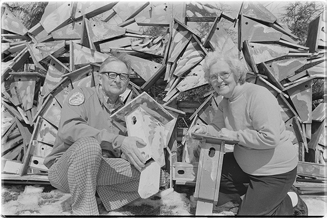 A man and a woman kneel on the ground, holding nest boxes. Behind them are more nest boxes in stacks that rise above their heads.