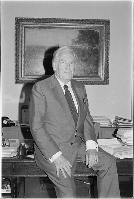 A man in a suit and tie sits on a desk in an office in front of a painting.