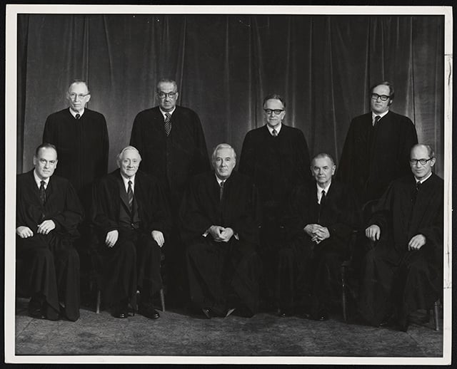 Nine US Supreme Court Justices sit and stand in two rows. They wear black robes.
