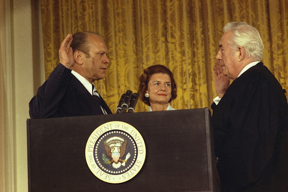 Three people stand behind a speaker's dais with the seal of the United States. At left, a man raises his right hand to mirror the man holding up his own right hand across from his. A woman stands in the middle, looking on.